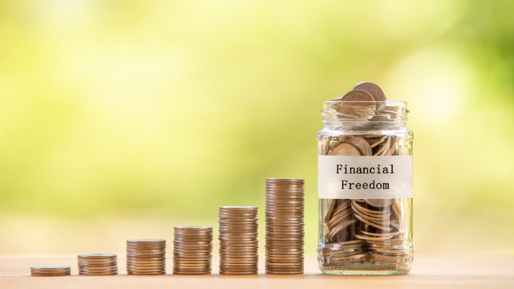 a glass jar with a text label stating "financial freedom" is filled with coins and is placed beside several piles of coins