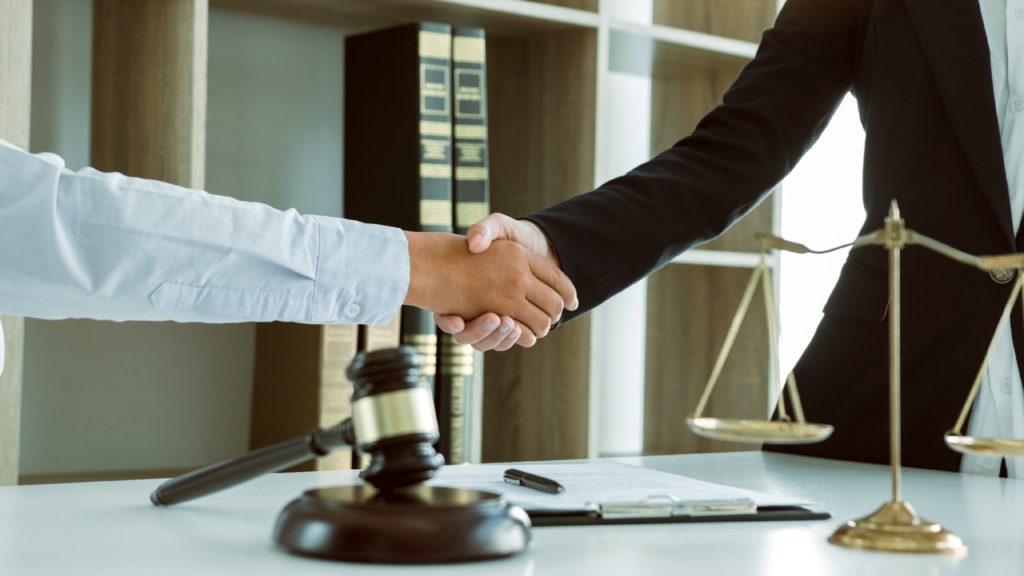 a lawyer shakes hands with a client over a desk featuring justice scales and a judge’s gavel