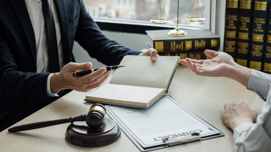 a lawyer speaks to a client as a gavel and paperwork sit between them