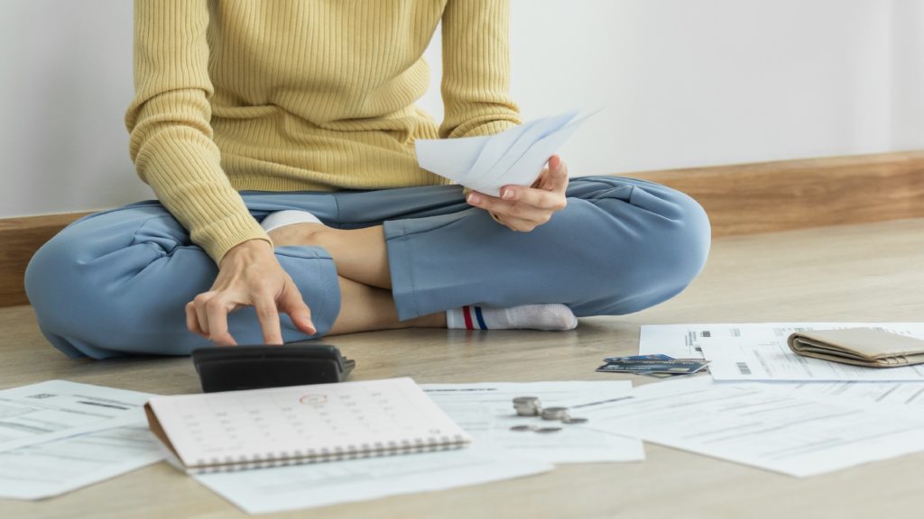 a person sits on the floor surrounded by documents as they type on a calculator