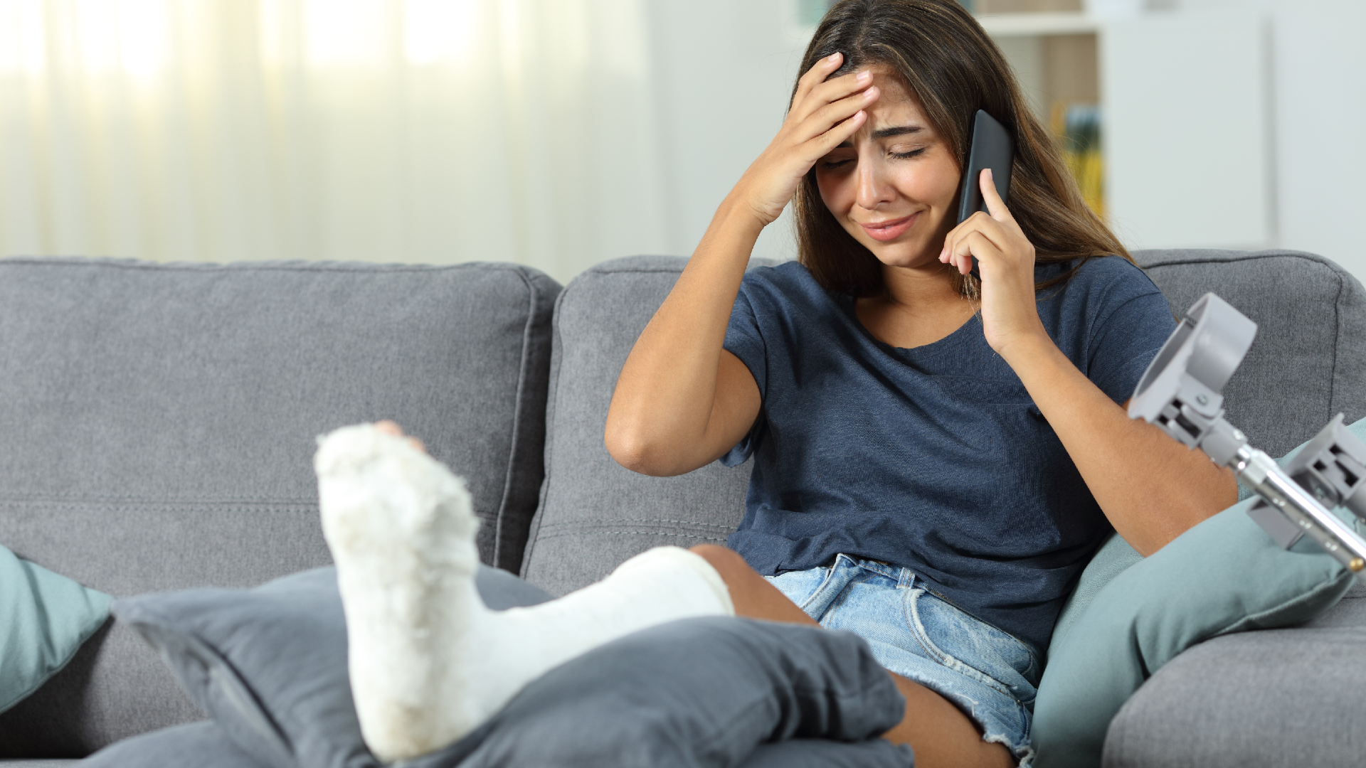 An injured woman with crutches and a cast on her leg talking on the phone while sitting on the couch and touching her head