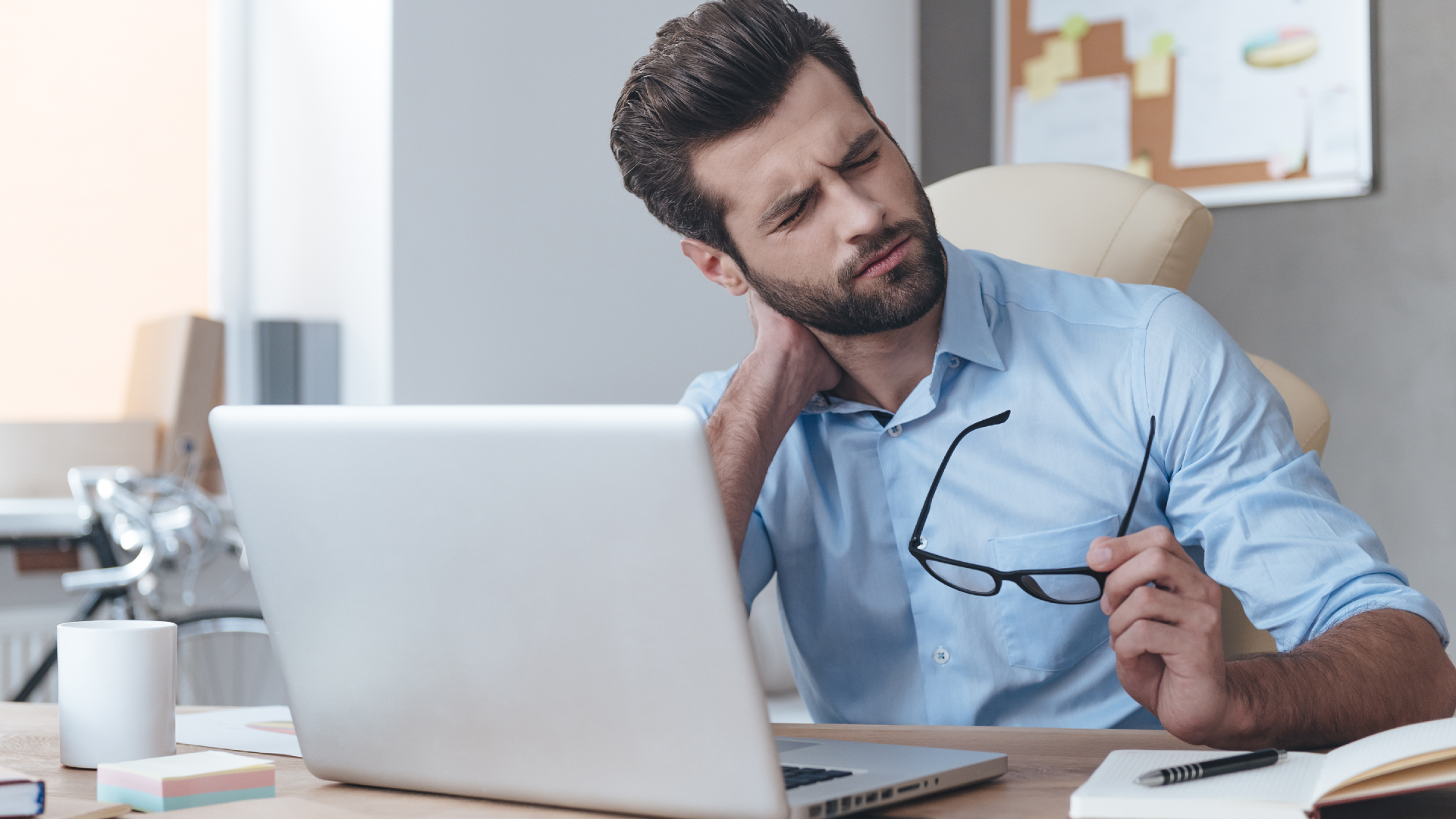 A man taking off his glasses and cracking his neck in front of a laptop computer