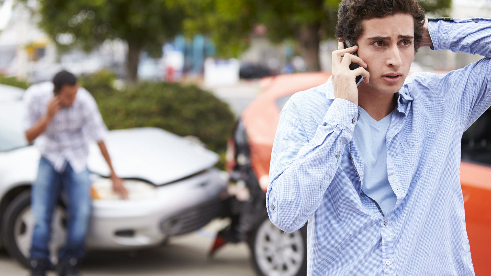 A man makes a phone call after a car accident while another man examines damage to his car in the background