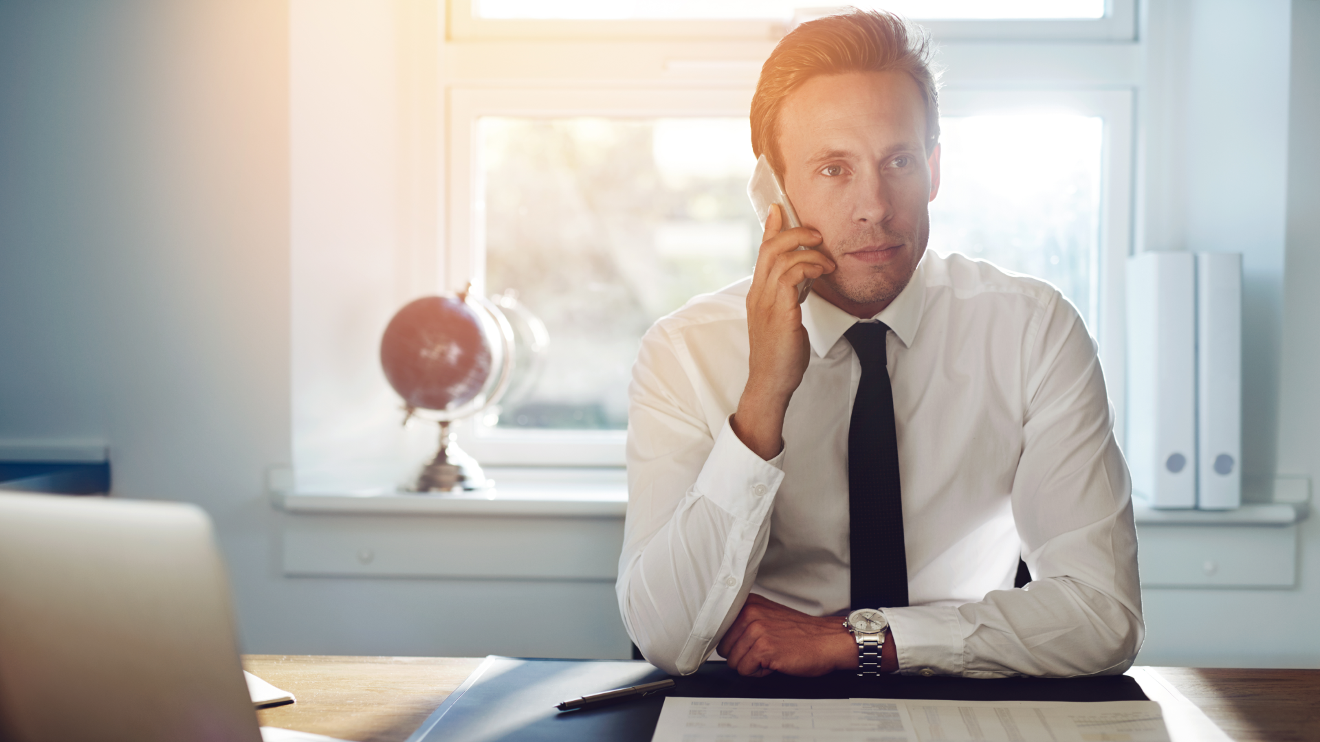 A man wearing a dress shirt and tie at his desk while talking on phone