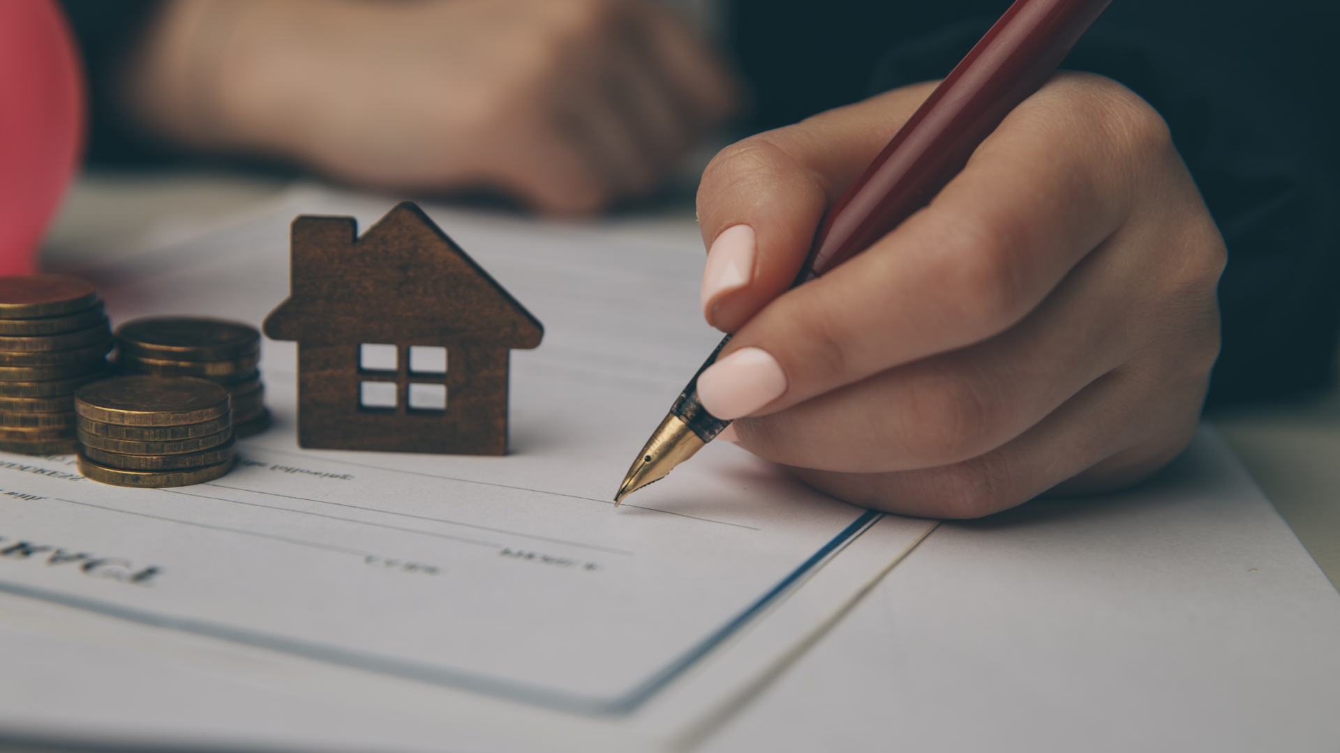 A person filling out paperwork with piles of coins and a model house on top