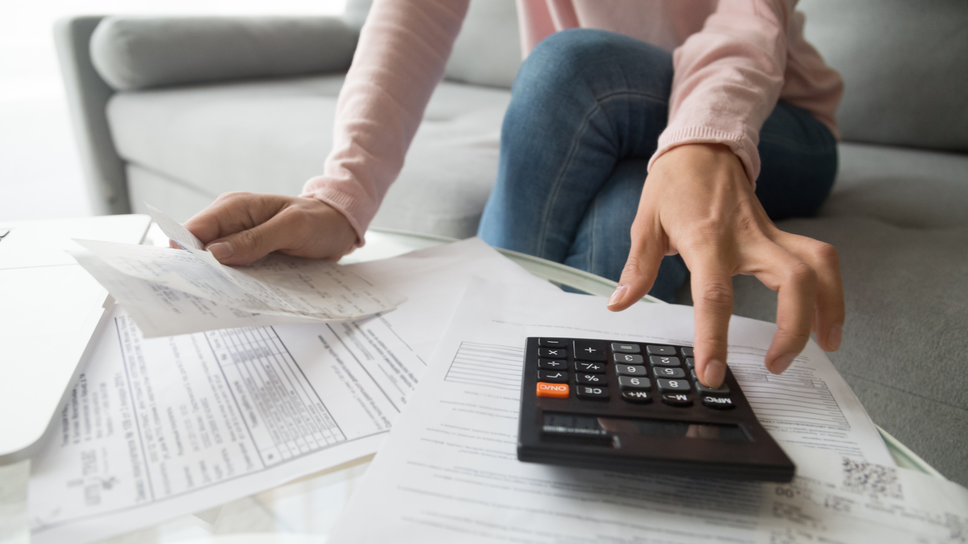 A woman sitting cross-legged on couch while looking over her finances and typing on a calculator